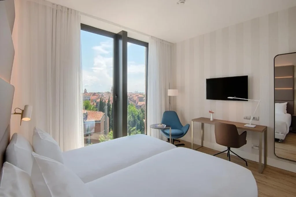 Bright and contemporary deluxe room featuring twin beds, floor-to-ceiling windows with Venice city views, a work desk, and stylish modern furnishings at Avani Rio Novo Venice Hotel in Venice, Italy.