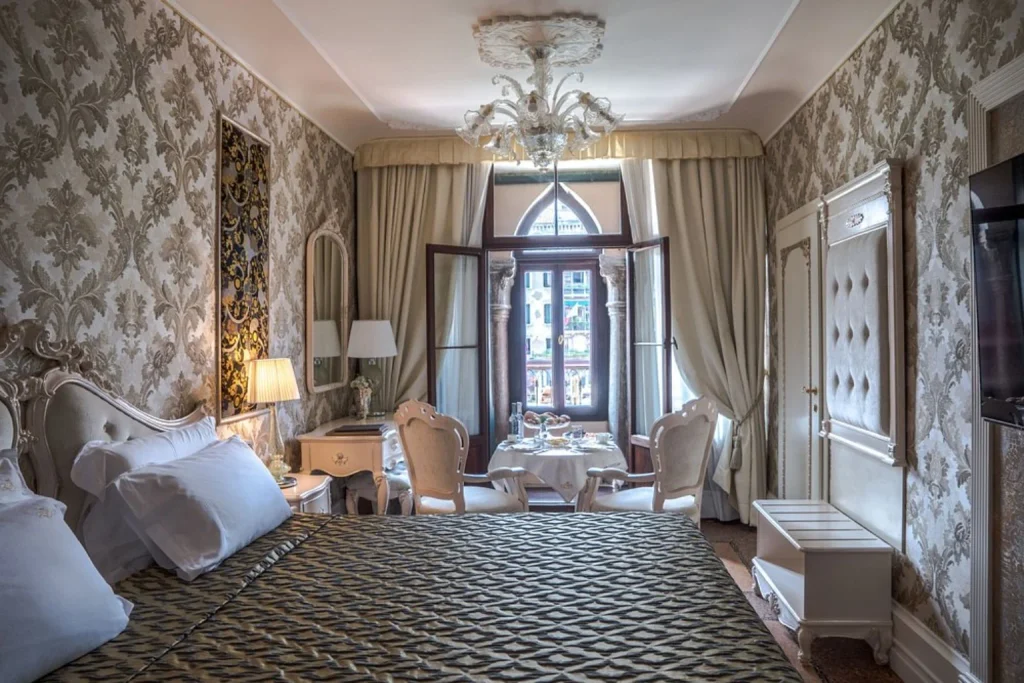 Luxurious bedroom featuring ornate damask décor, a crystal chandelier, elegant period-style furnishings and tall windows with a canal view – Ego’ Boutique Hotel The Silk Road, Venice.