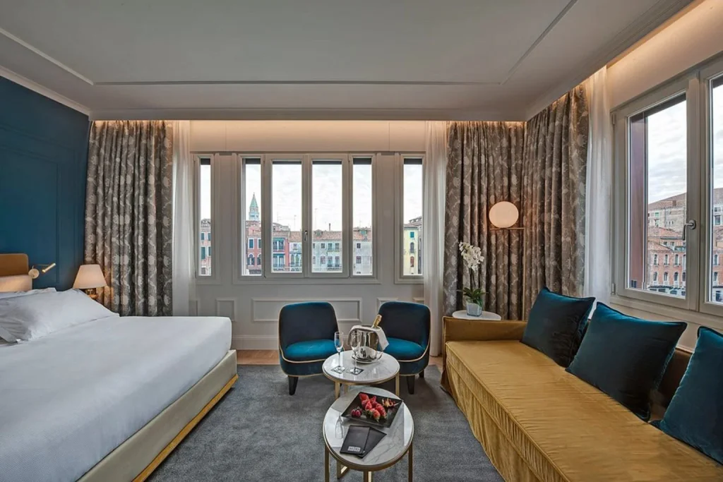 Luxury hotel bedroom with elegant seating, floor-to-ceiling windows and panoramic views of historic Venetian buildings – H10 Palazzo Canova, Venice