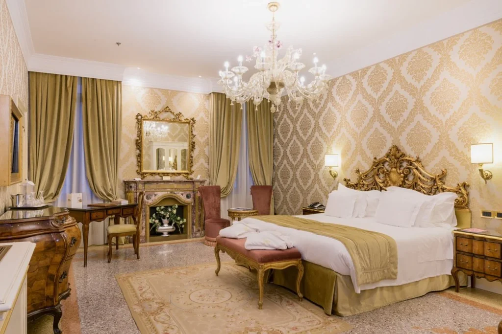 Luxurious Venetian bedroom with an ornate carved bed, crystal chandelier, damask wallpaper and antique-style furnishings – Hotel Ai Reali – Small Luxury Hotels of the World, Venice