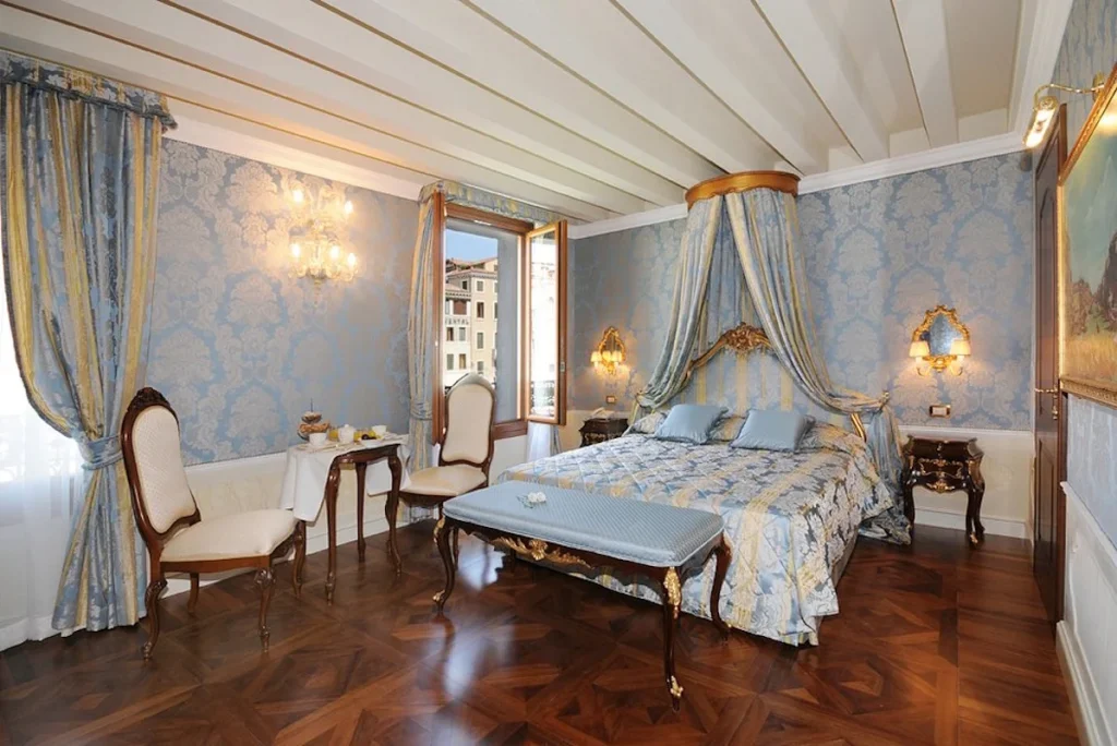 Elegant deluxe room with a canopy bed, blue Venetian décor, crystal chandelier, and hardwood floors at Hotel Canal Grande in Venice, Italy.