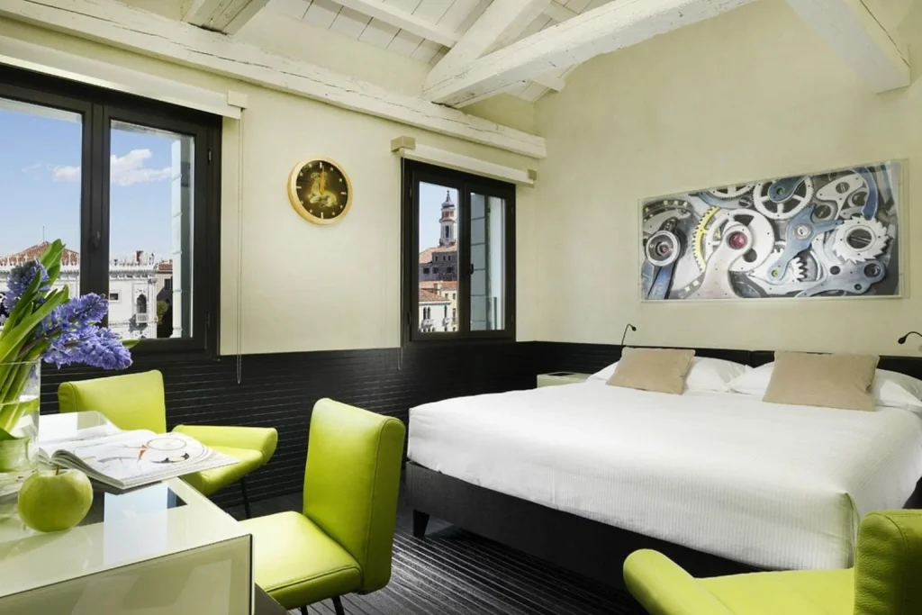 Stylish bedroom with exposed white beams, contemporary décor and large windows offering views of historic rooftops and a bell tower – Hotel L’Orologio Venezia, Venice.