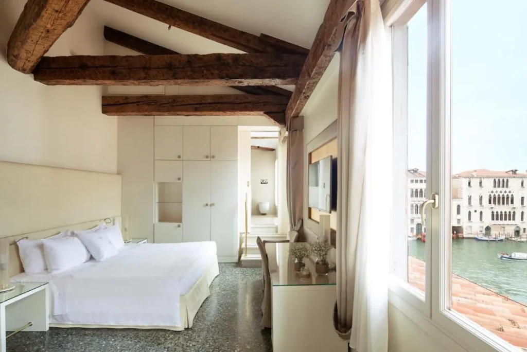 Grand Canal view room with exposed wooden beams, modern furnishings, and large windows overlooking Venice at Palazzo Barocci in Venice, Italy.