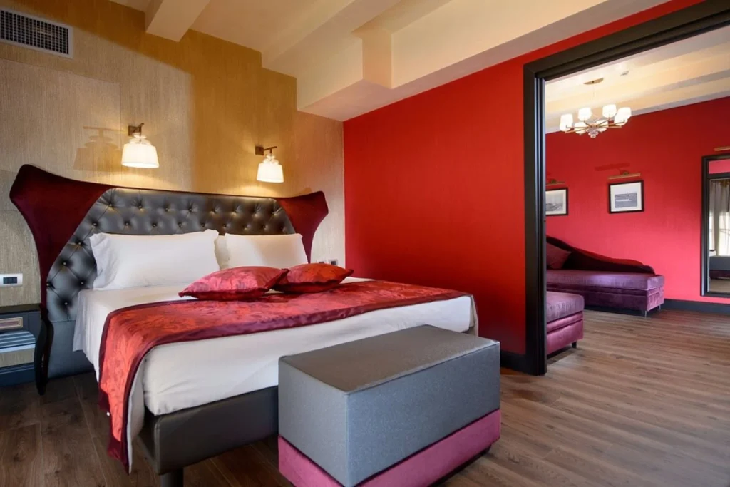 Spacious luxury suite featuring a statement upholstered bed, rich red décor and a separate sitting area, Palazzo Veneziano, Venice.