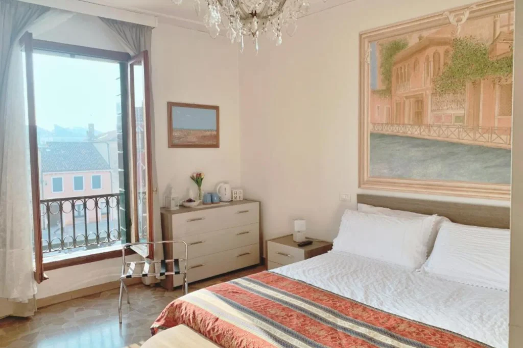 Bright Venetian bedroom with chandelier, decorative artwork and an open balcony overlooking the city – Venezia Canal View, Venice